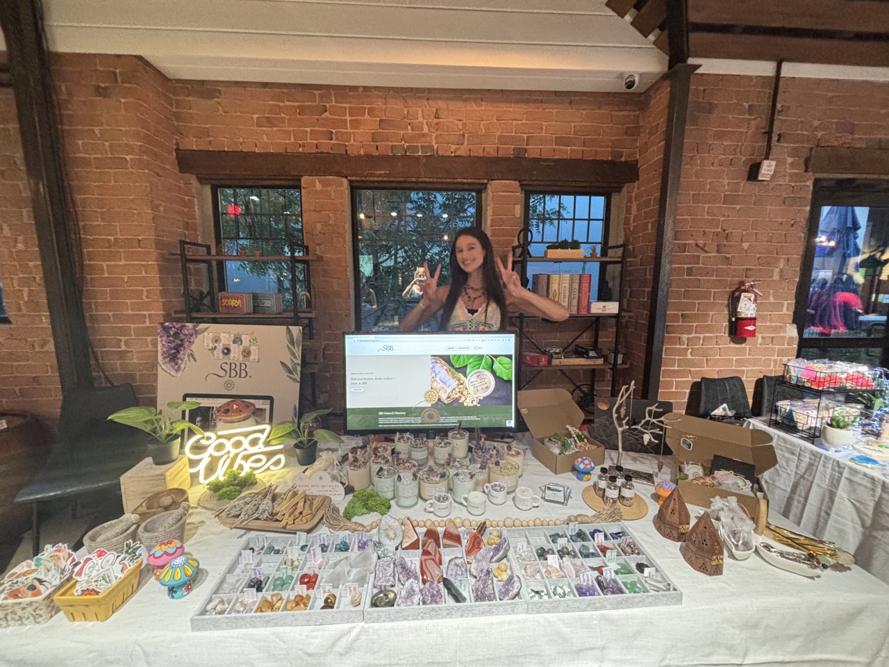 Liz Holding Up Two Peace Signs Standing Behind Booth at Pop Up Shop Showing Holistic Product Offerings